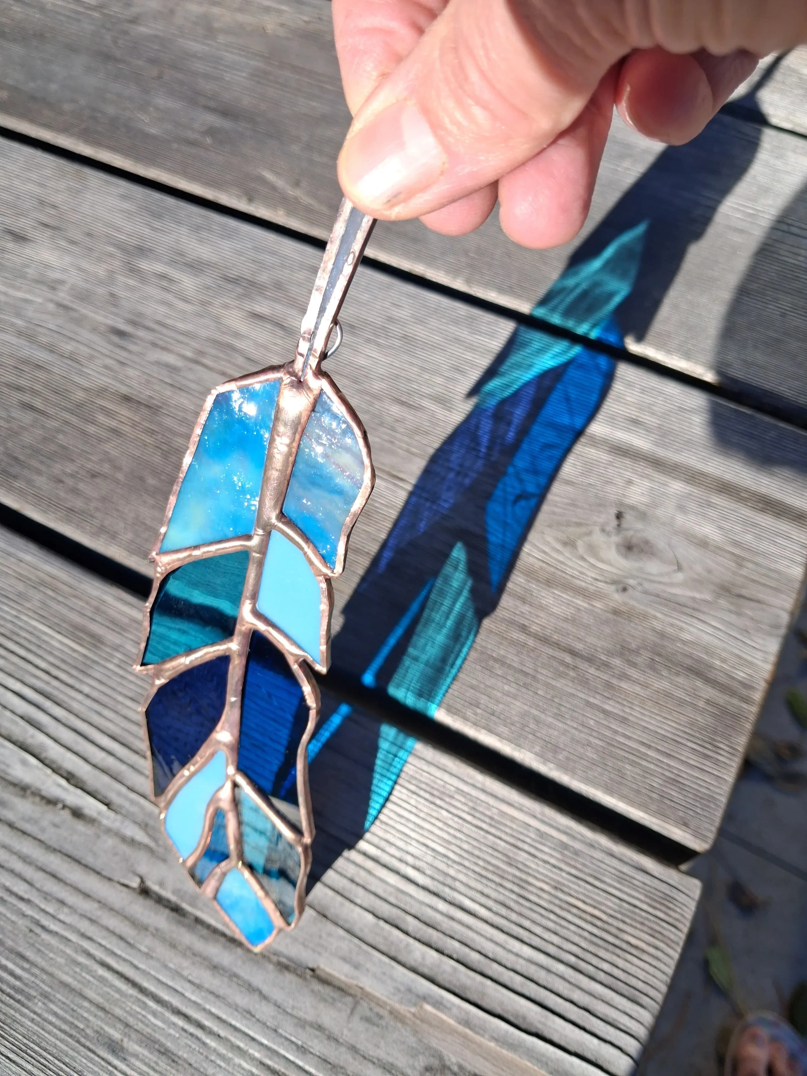 Blue stained glass feather plant stake casting colorful light on wooden deck