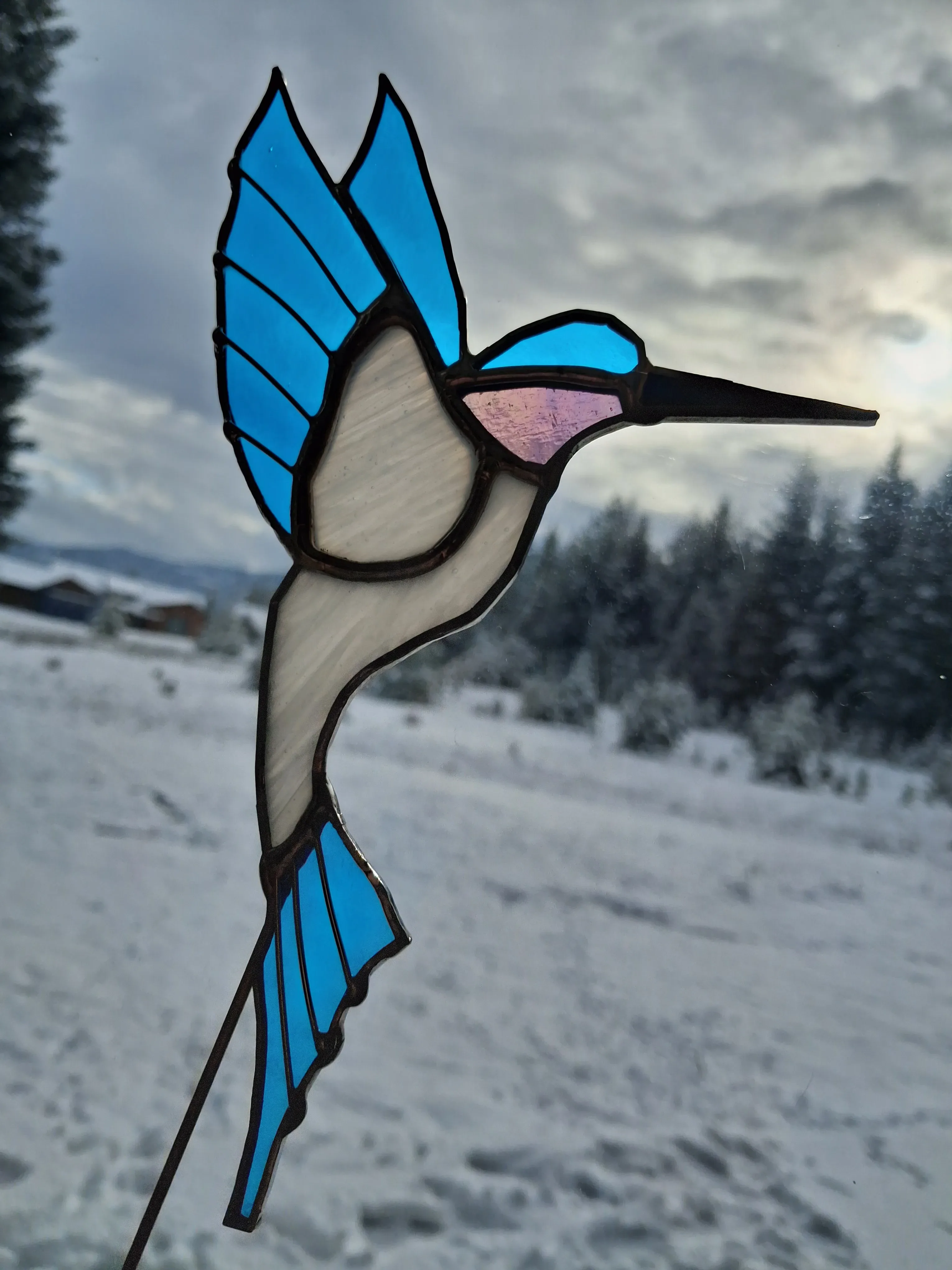 Blue and white stained glass hummingbird suncatcher with pink throat against snowy Idaho landscape