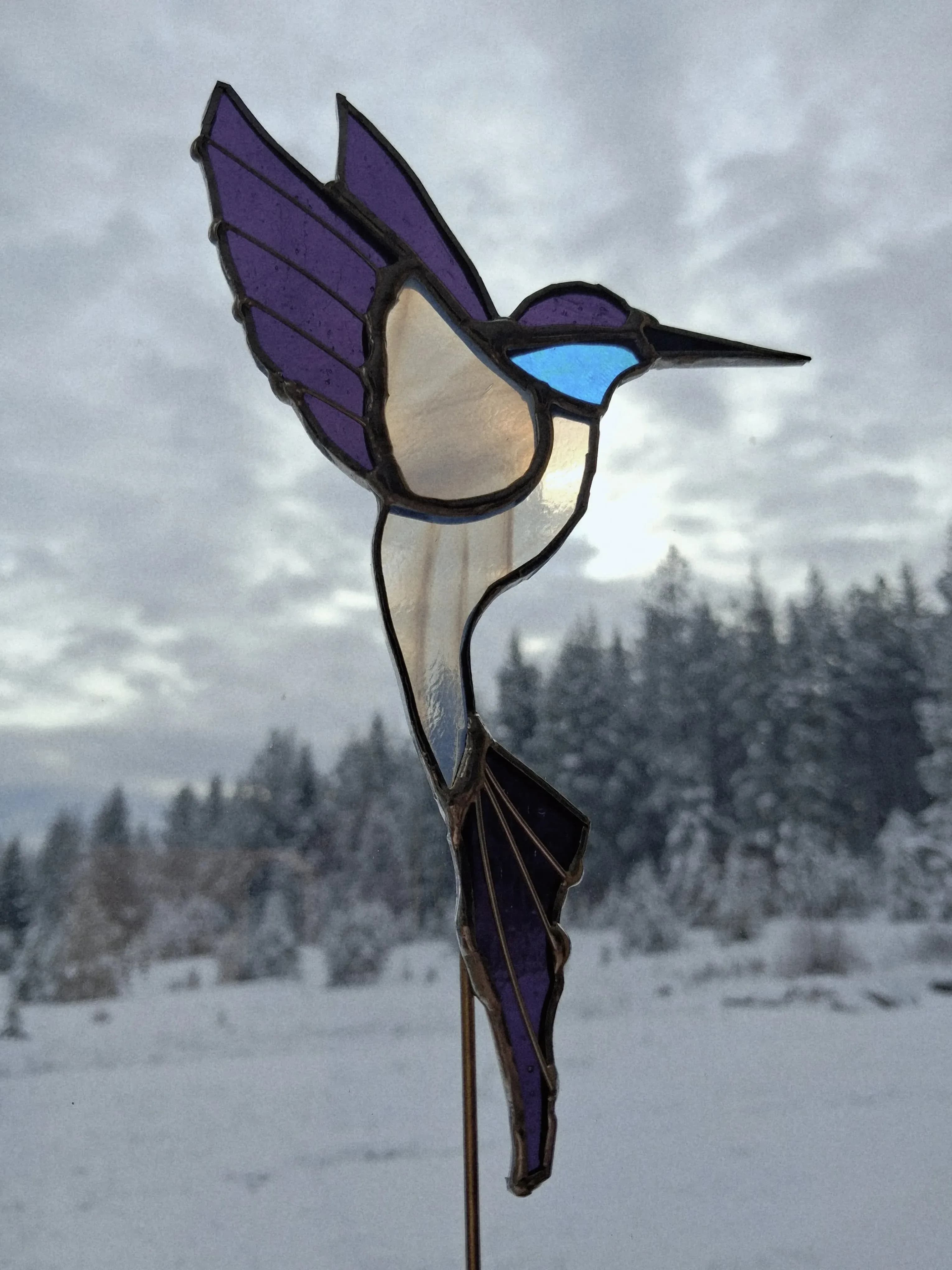 Purple and blue stained glass hummingbird suncatcher with iridescent accents against snowy trees
